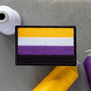 Peut inclure: Un écusson brodé jaune, blanc, violet et noir avec les couleurs disposées en bandes horizontales. L'écusson est sur une surface grise avec des bobines de fil jaune, blanc et violet.