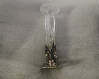 May include: A clear glass vase with a fluted, vertical ribbed design. The base is adorned with a metallic gold-toned decorative element featuring dark brown, curved leaf-like accents and small, light purple and yellow floral details.