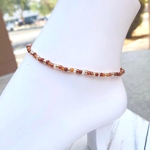 May include: A delicate ankle bracelet made with small brown, orange, and gold beads.