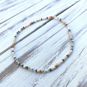 May include: Delicate beaded necklace featuring a mix of small, colorful beads. The necklace includes beads in shades of teal, gold, peach, and white. The necklace is displayed on a white wooden surface, showcasing its intricate design and color palette.