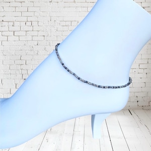 May include: A delicate beaded anklet featuring small, dark gray and silver beads strung together. The anklet is displayed on a light blue foot form, showcasing its design and fit. The anklet is a simple yet elegant piece of jewelry.
