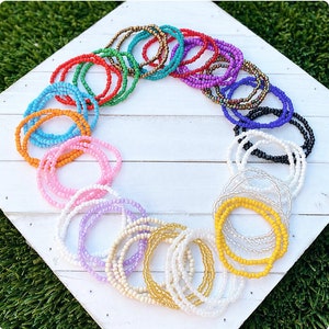 May include: A collection of colorful beaded bracelets in various shades of pink, blue, yellow, green, purple, orange, and black. The bracelets are arranged in a circle on a white wooden surface.
