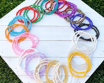 May include: A collection of colorful beaded bracelets in various shades of pink, blue, yellow, green, purple, orange, and black. The bracelets are arranged in a circle on a white wooden surface.