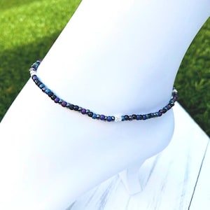 May include: A delicate anklet made with small iridescent beads in shades of blue, purple, and black. The anklet is accented with a single silver bead.
