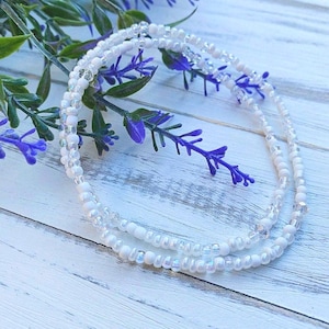 May include: Two strands of white beaded jewelry with iridescent accents. The beads are small and round, and the jewelry is lying on a white wooden surface.