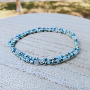 May include: A delicate, multi-layered bracelet made of small, iridescent blue and white beads. The bracelet is wrapped around a wooden surface.