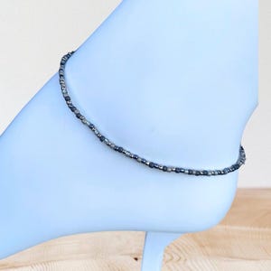 May include: Ankle bracelet with small, dark gray and silver beads. The beaded jewelry is worn around the ankle. The anklet is on a light blue surface.