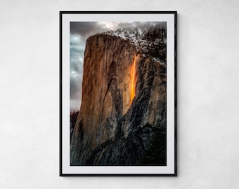 May include: A photograph of a large rock formation with a waterfall cascading down its side. The waterfall is illuminated by the setting sun, creating a fiery orange glow. The rock face is a mix of gray and brown, with some snow visible at the top.