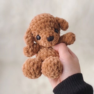 May include: A brown crocheted plush toy poodle with black eyes and a black nose. The toy is being held in a hand.