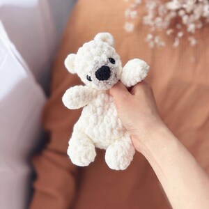 May include: A small, white crocheted teddy bear with a black nose and eyes. The bear has a textured, fluffy appearance and is held in a person's hand. The bear's ears and paws are also white.