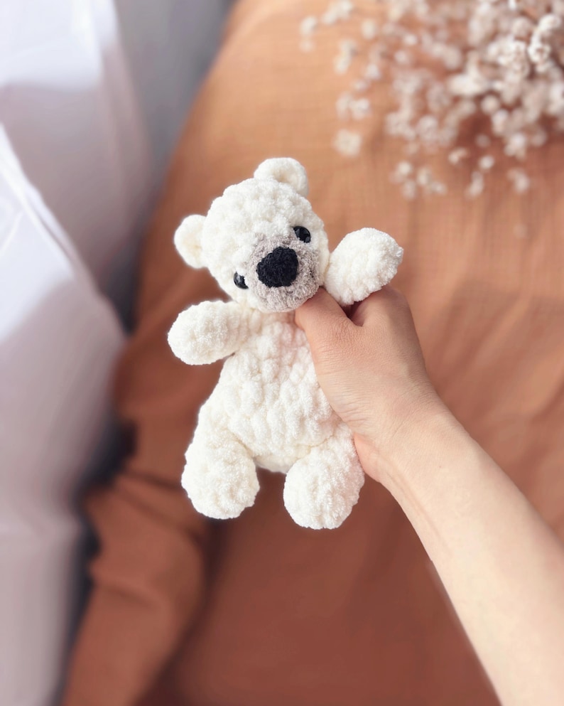 May include: A small, white crocheted teddy bear with a black nose and eyes. The bear has a textured, fluffy appearance and is held in a person's hand. The bear's ears and paws are also white.