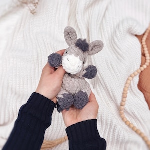 May include: A small, handmade crochet donkey plush toy crafted from soft, textured gray yarn. The donkey features a white muzzle, dark gray hooves, a small tuft of dark gray hair on its head, and embroidered closed eyes, creating a gentle appearance.
