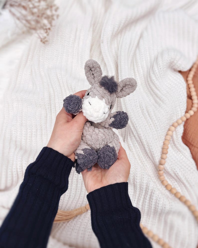 May include: A small, handmade crochet donkey plush toy crafted from soft, textured gray yarn. The donkey features a white muzzle, dark gray hooves, a small tuft of dark gray hair on its head, and embroidered closed eyes, creating a gentle appearance.