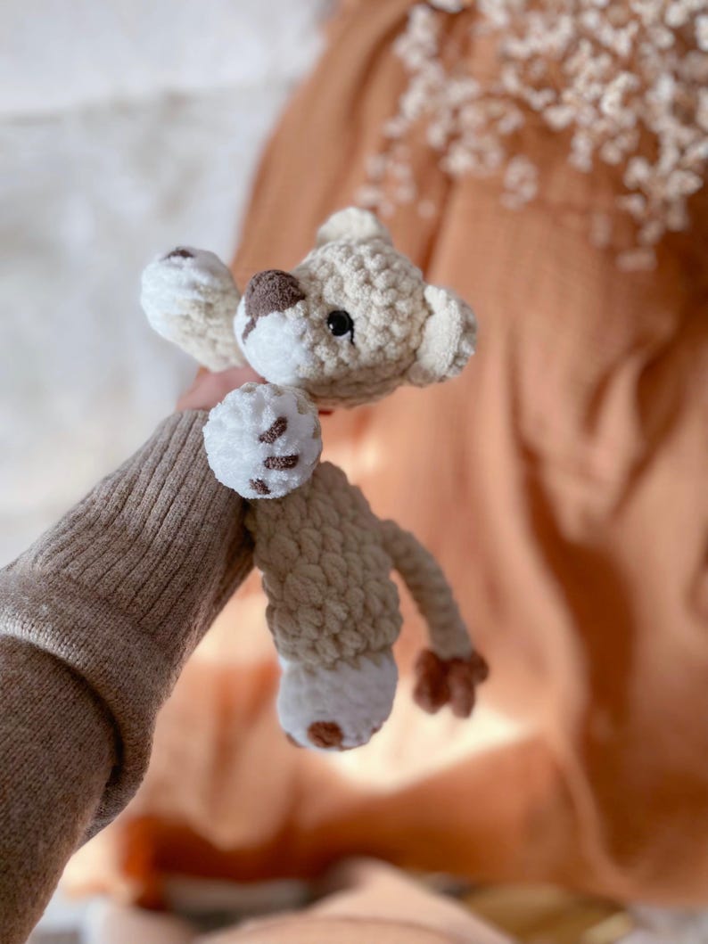 May include: A crocheted stuffed animal toy featuring a light beige body with white accents on the paws and face. The toy has a dark brown nose, small black eyes, and a long tail with a dark brown tip, crafted from a soft, textured yarn material.