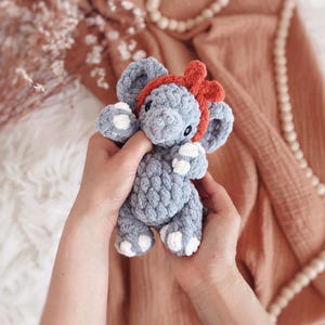 May include: A handmade grey crochet elephant toy with white detailing on its feet and ears, and a red bow headband. The plush toy is held in two hands, highlighting its soft texture and detailed craftsmanship. A charming and cuddly children's toy.
