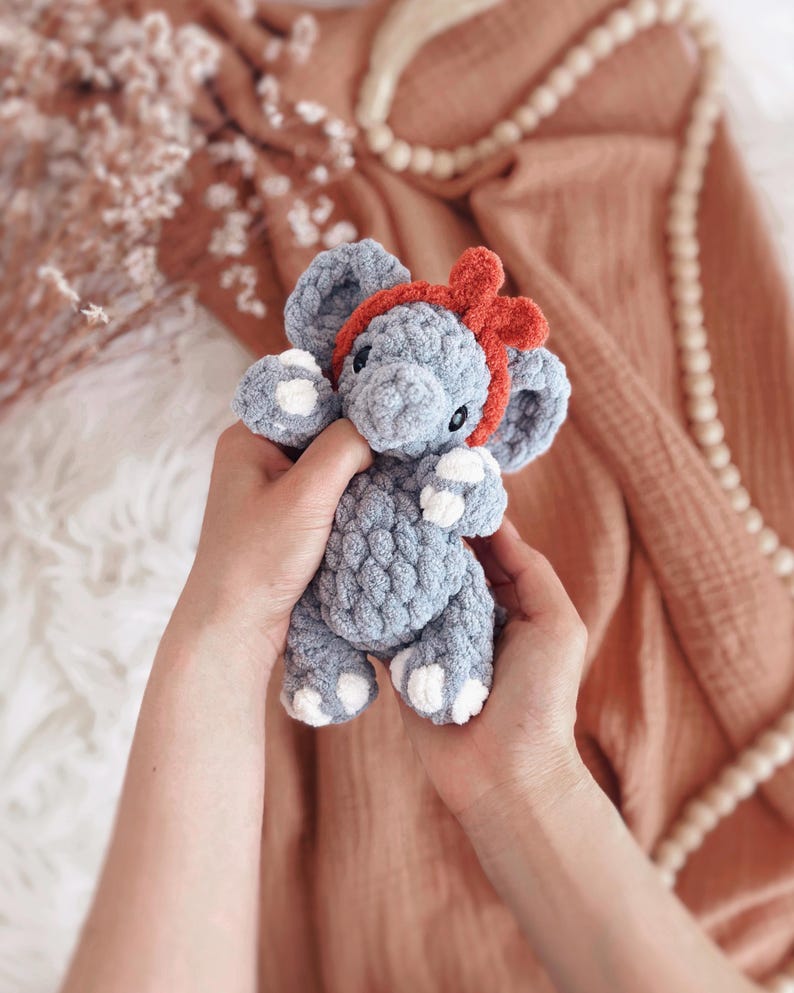 May include: A handmade grey crochet elephant toy with white detailing on its feet and ears, and a red bow headband. The plush toy is held in two hands, highlighting its soft texture and detailed craftsmanship. A charming and cuddly children's toy.