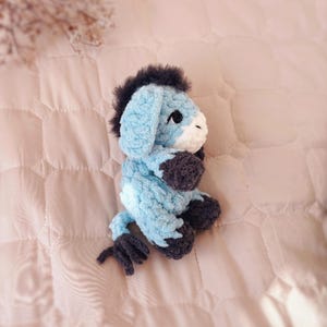 May include: A small, handmade crochet donkey plush toy crafted from soft, light blue yarn. It features a dark gray mane, tail, and hooves, with a white muzzle and black safety eyes. The donkey is depicted in a seated position, showcasing its textured, fuzzy yarn construction and detailed stitching throughout the body.