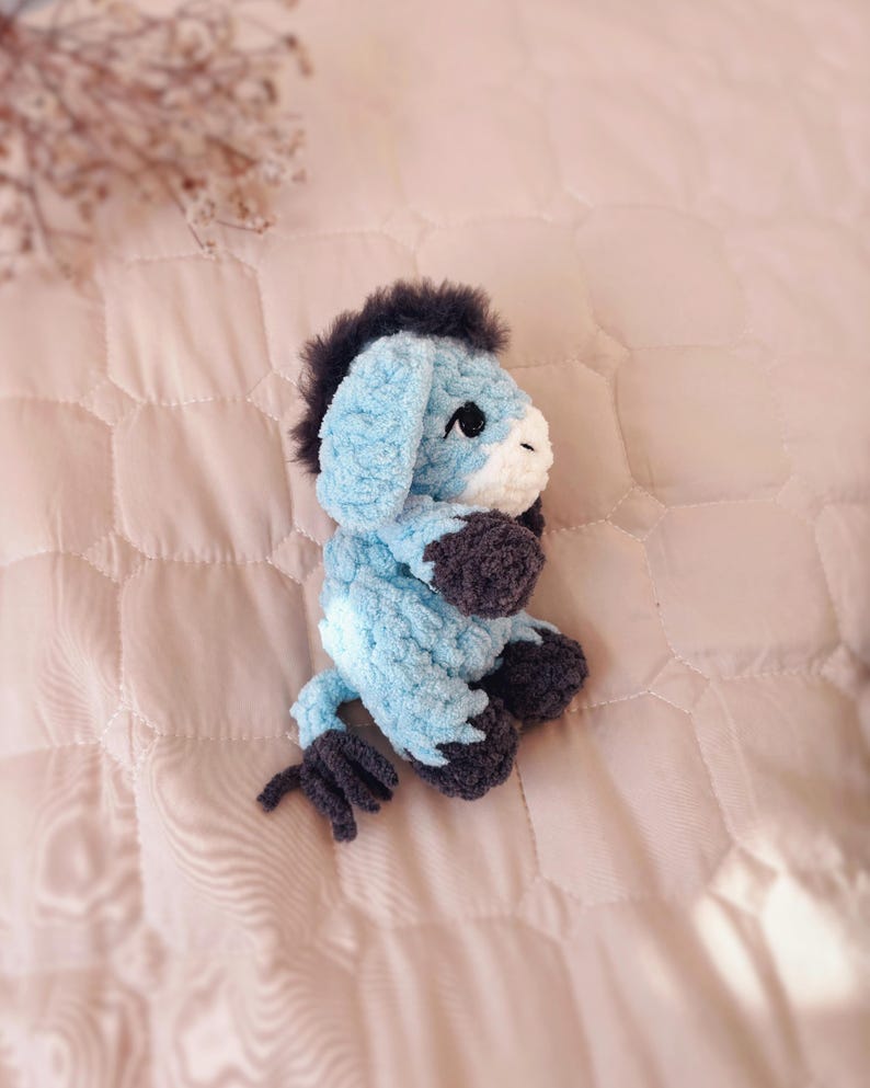 May include: A small, handmade crochet donkey plush toy crafted from soft, light blue yarn. It features a dark gray mane, tail, and hooves, with a white muzzle and black safety eyes. The donkey is depicted in a seated position, showcasing its textured, fuzzy yarn construction and detailed stitching throughout the body.