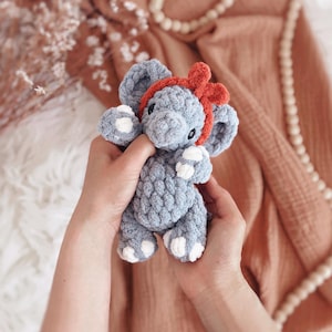 May include: A handmade grey crochet elephant toy with white detailing on its feet and ears, and a red bow headband. The plush toy is held in two hands, highlighting its soft texture and detailed craftsmanship. A charming and cuddly children's toy.