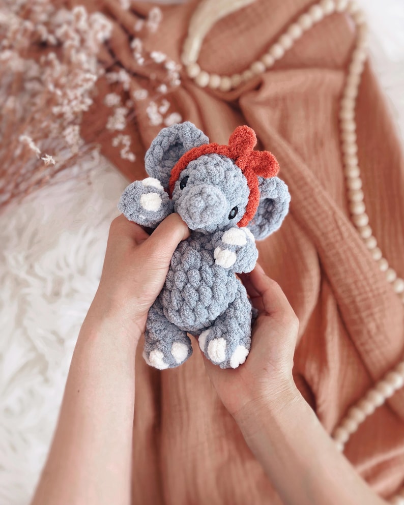 May include: A handmade grey crochet elephant toy with white detailing on its feet and ears, and a red bow headband. The plush toy is held in two hands, highlighting its soft texture and detailed craftsmanship. A charming and cuddly children's toy.