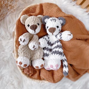 May include: Two plush crochet toys, one a light beige bear and the other a white tiger with grey stripes. Both toys feature soft, textured yarn, dark brown embroidered noses, and white paw pads with brown or pink accents on their feet.