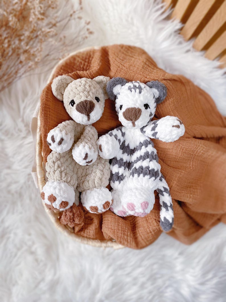 May include: Two plush crochet toys, one a light beige bear and the other a white tiger with grey stripes. Both toys feature soft, textured yarn, dark brown embroidered noses, and white paw pads with brown or pink accents on their feet.