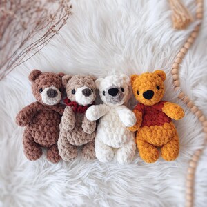 May include: Four handmade crochet teddy bears in various colors: brown, tan, white, and orange. Each bear has a unique expression and design, with details like a red scarf and black eyes. The bears are arranged on a fluffy white surface.
