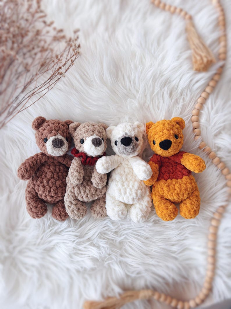 May include: Four handmade crochet teddy bears in various colors: brown, tan, white, and orange. Each bear has a unique expression and design, with details like a red scarf and black eyes. The bears are arranged on a fluffy white surface.