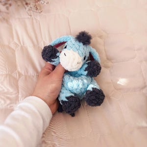 May include: A small, light blue crocheted donkey plush toy with a white muzzle, dark gray hooves, and a dark gray tuft of hair on its head. The stuffed animal features long, floppy ears and a soft, textured yarn construction throughout its body.
