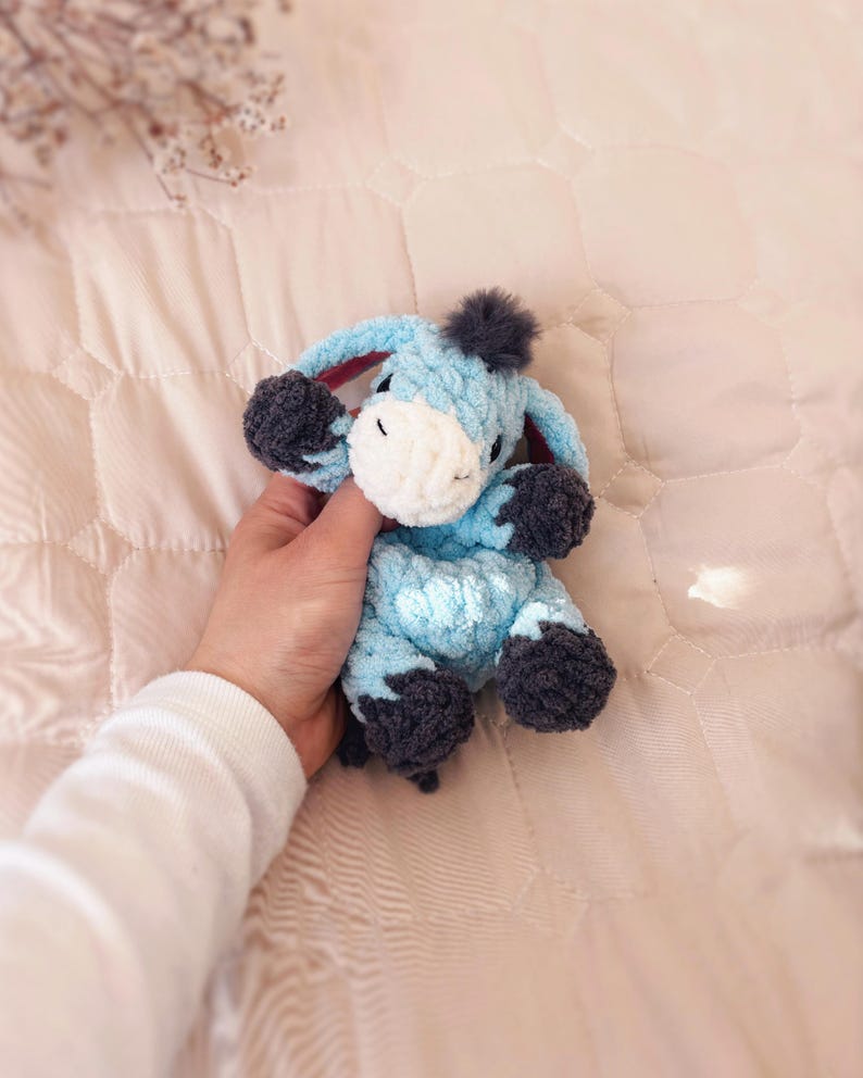 May include: A small, light blue crocheted donkey plush toy with a white muzzle, dark gray hooves, and a dark gray tuft of hair on its head. The stuffed animal features long, floppy ears and a soft, textured yarn construction throughout its body.
