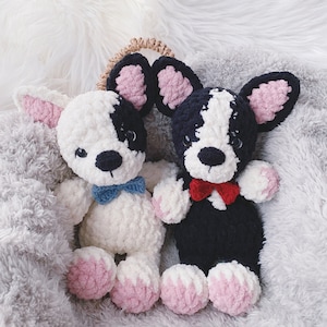 May include: Two crocheted stuffed dog toys, one white with blue bow tie and one black with red bow tie, sitting on a fluffy gray blanket.