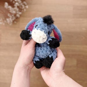 May include: A small, handmade crochet donkey plush toy made from soft, textured blue yarn. It features a white muzzle, black hooves, a black tuft of hair on its head, and large ears with pink inner lining. The donkey has embroidered eyes.
