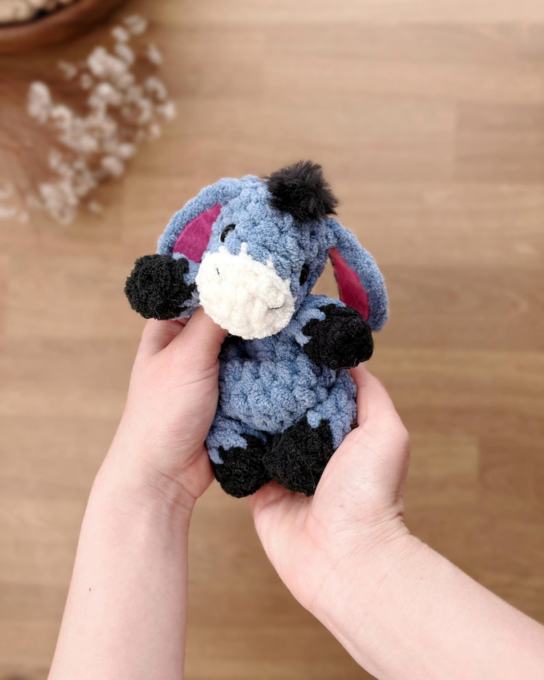 May include: A small, handmade crochet donkey plush toy made from soft, textured blue yarn. It features a white muzzle, black hooves, a black tuft of hair on its head, and large ears with pink inner lining. The donkey has embroidered eyes.