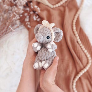 May include: A handmade grey crochet elephant toy with white detailing on its feet and ears. The elephant has a light beige headband and a small bow. The toy is held in a person's hands, with a blurred background of brown fabric and decorative beads.