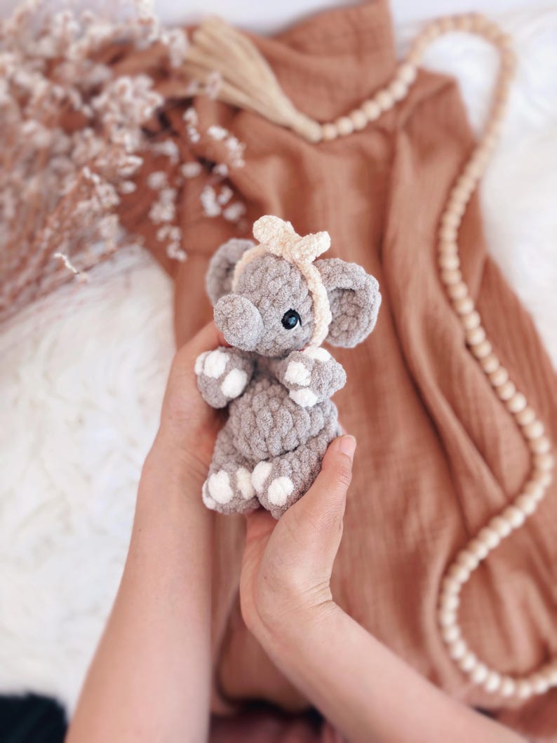 May include: A handmade grey crochet elephant toy with white detailing on its feet and ears. The elephant has a light beige headband and a small bow. The toy is held in a person's hands, with a blurred background of brown fabric and decorative beads.