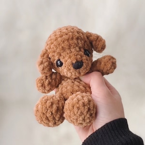 May include: A brown crocheted plush toy poodle with black eyes and a black nose. The toy is being held in a hand wearing a black sweater.
