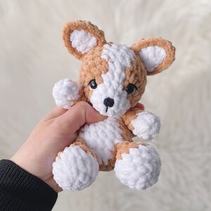 May include: A crocheted stuffed dog toy with brown and white fur. The dog is sitting in a hand.