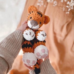 May include: A crocheted tiger plush toy featuring orange yarn with black stripes, a white muzzle, and white paws with pink embroidered pads. The stuffed animal has small rounded ears, dark eyes, and a soft, textured yarn construction throughout.