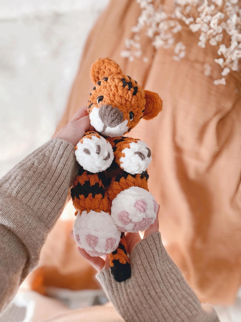 May include: A crocheted tiger plush toy featuring orange yarn with black stripes, a white muzzle, and white paws with pink embroidered pads. The stuffed animal has small rounded ears, dark eyes, and a soft, textured yarn construction throughout.