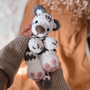 May include: A plush tiger toy made of soft white and dark grey yarn. The stuffed animal features a rounded head with dark grey ears, a brown nose, and embroidered eyes. Its paws are white with light pink stitched pads on the bottom of the feet.