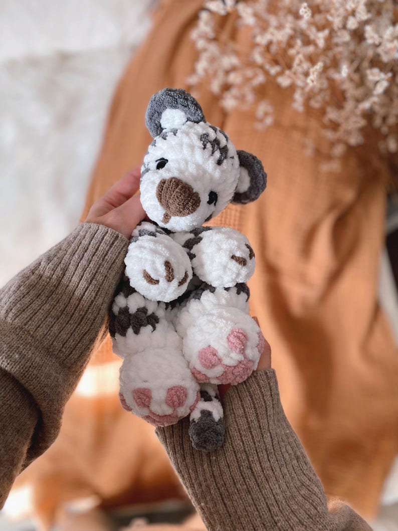 May include: A plush tiger toy made of soft white and dark grey yarn. The stuffed animal features a rounded head with dark grey ears, a brown nose, and embroidered eyes. Its paws are white with light pink stitched pads on the bottom of the feet.