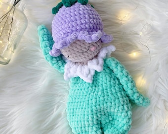 Puede incluir: Una muñeca de crochet con un sombrero de flor morado, un cuello blanco y un cuerpo turquesa. La muñeca está tumbada sobre una superficie blanca y esponjosa.