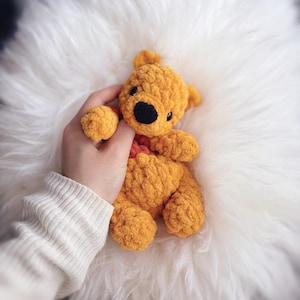 May include: A handmade, golden yellow crochet teddy bear with a black nose and eyes. The bear has a red heart-shaped detail and is resting on a fluffy white surface. The bear is being held by a person wearing a white sweater.