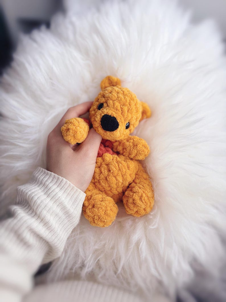May include: A handmade, golden yellow crochet teddy bear with a black nose and eyes. The bear has a red heart-shaped detail and is resting on a fluffy white surface. The bear is being held by a person wearing a white sweater.