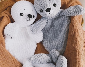 Op de afbeelding: Twee gehaakte pluche zeehondenspeelgoedjes, een wit en een grijs, zitten in een bruine mand. De speelgoedjes hebben zwarte ogen en neuzen. De tekst "English | PDF file" is zichtbaar in het midden onderaan de afbeelding.
