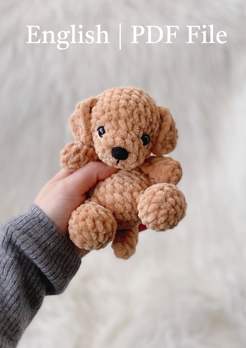 May include: A crocheted plush toy dog in a light brown color. The dog has black eyes and a black nose. The dog is being held in a person's hand.
