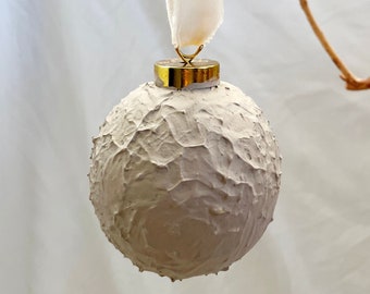 May include: A white, textured, round ornament with a gold top and a white ribbon hanging from it.