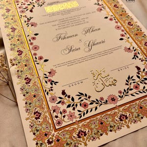 Pu&ograve; includere: Un invito di nozze con un design floreale dorato e rosa. Il testo recita "This agreement made on the 12th day of April 2005 corresponding to the 13th day of Shawwal, 1446 AH in the city of Edison, NJ is to certify the marriage of Fehman & Sara Ghauri".
