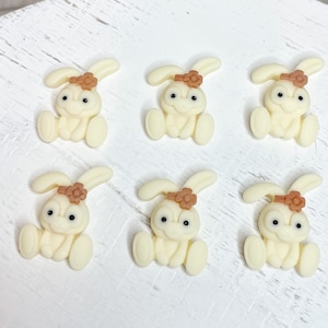 以下が含まれることがあります： 茶色の花のアクセントが付いた、クリーム色のウサギの置物6つ。各ウサギは黒い目を持ち、直立しています。これらの置物は白い表面に配置されており、工芸品や装飾をテーマにしていることを示唆しています。
