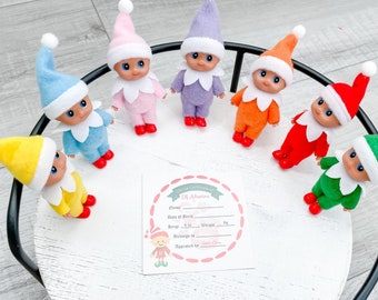May include: Seven colorful elf dolls with matching hats and outfits are arranged on a white, round tray. Each doll has a different color scheme, including yellow, blue, pink, purple, orange, red, and green. A card with elf adoption details is also present.